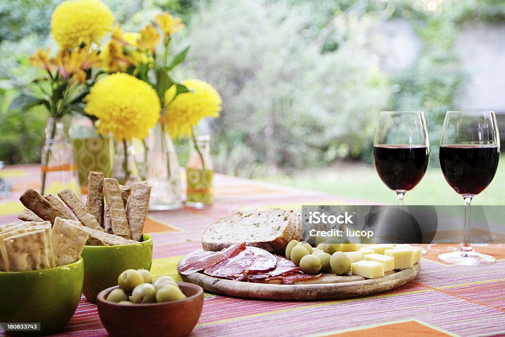 Party table with yellow flowers, wine, buffet at garden A DSLR photo, with a eye level perspective, of a table setting with food, yellow flowers and red decor at a lush garden in a beautiful sunny afternoon. The table is dressed with a colourful printed design tablecloth. Two glasses of red wine stands on the right. A cold buffet food is on the foreground featuring  breadsticks, olives, cheese, prosciutto and sliced bread. A the background is a centrepiece arrangement with yellow chrysanthemum flowers in vintage glass bottles. The garden is defocused at the background. Copy space at the top. Cheese Stock Photo