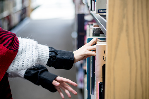 In the silent dance of fingertips, a gender non-conforming teen delicately selects a book from the library shelf. This close-up moment unveils the power of choice and the profound connection between personal identity and the narratives we embrace