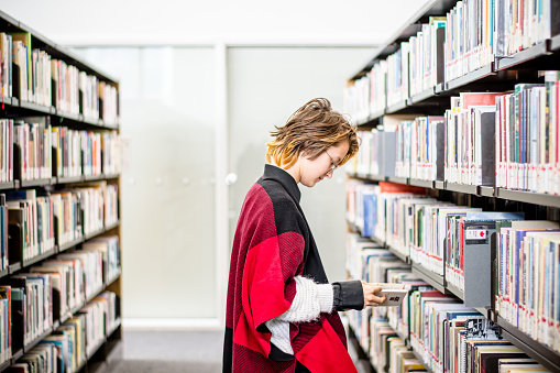 In the quiet corners of the public library, a 13-year-old gender non-conforming teen confidently navigates shelves, finding solace and inspiration among the bound worlds of literature. Their short pixie haircut and trendy autumn attire speak volumes about the freedom to be true to oneself in every space.