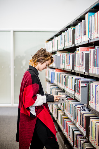 In the quiet corners of the public library, a 13-year-old gender non-conforming teen confidently navigates shelves, finding solace and inspiration among the bound worlds of literature. Their short pixie haircut and trendy autumn attire speak volumes about the freedom to be true to oneself in every space.