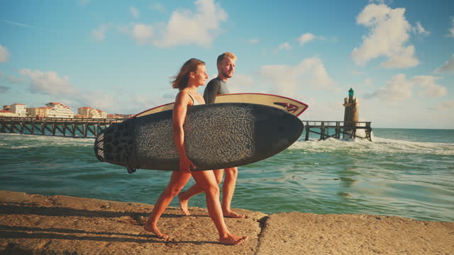 SLO MO Surfer couple walking with surfboards along sunny ocean