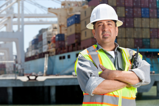 A dock worker with a clipboard and pen at a port with a cargo ship and crane in the background.