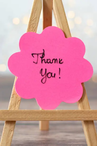 Thank you text handwritten on pink sticky note in flower shape with bokeh background. Close-up, vertical photo. Concept of thanksgiving, gratitude, and acknowledgment.