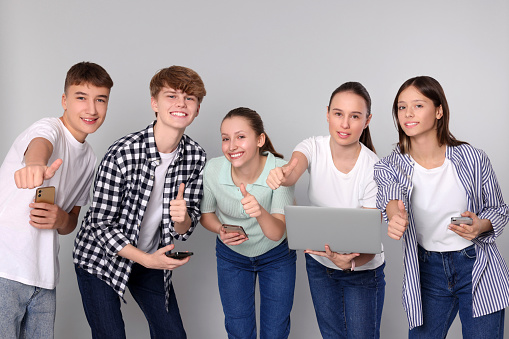 Group of happy teenagers with smartphones and laptop showing thumbs up on light grey background