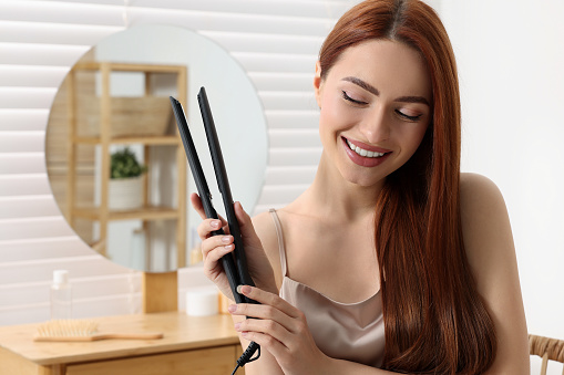 Beautiful woman with hair iron in room, space for text