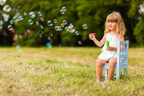 Beautiful little girl sitting outdoors blowing bubbles on a summer day. 