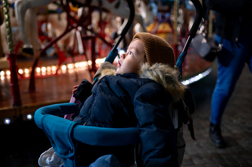 Cute little boy siting in a baby carriage and looking at the carousel, horses and lights at amusement park in Italy. Exciting and fun winter cold evening for baby on a trip to Italy
