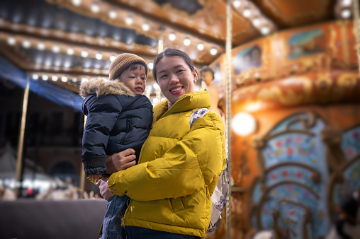 Cute little boy with a knitted beanie hat is enjoying with his mother on carousel in an Italian amusement park. Smiling young mom is standing and holding her little son in her arms. The photo was taken on a cold winter day during a trip to Italy