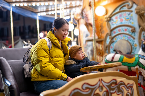 Cute little boy with a knitted beanie hat is sitting with his mother on carousel in an Italian amusement park. Boy holding a wooden steering wheel and looking around. The photo was taken on a cold winter day during a trip to Italy. The boy's father is waiting for them to finish the ride