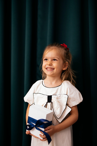 Close up shot of a happy little girl in a white dress with a big bow, standing and holding a gift box in her hands. She is smiling and looking away.