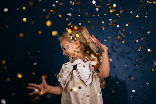 Low angle view of a smiling girl in elegant dress having fun while confetti falling down during a Christmas celebration.