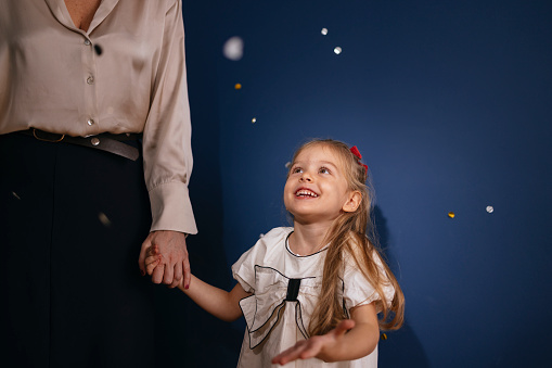 A smiling daughter holding her mother hand, looking away and standing together while confetti falling down during a New Year celebration.