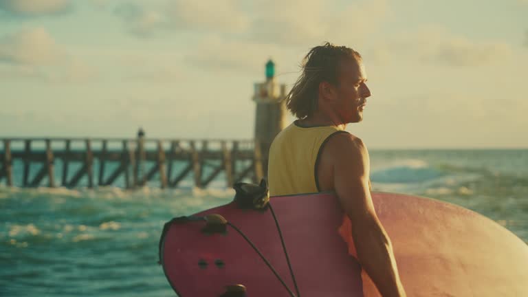 SLOW MOTION Male surfer with surfboard walking along sunny ocean pier