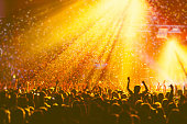 A crowded concert hall with scene stage orange and yellow lights, rock show performance, with people silhouette, colourful confetti explosion fired on dance floor air during a concert festival