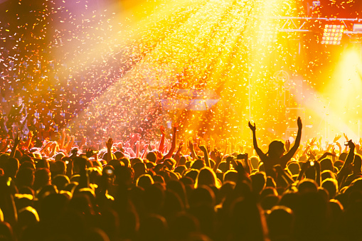 A crowded concert hall with scene stage orange and yellow lights, rock show performance, with people silhouette, colourful confetti explosion fired on dance floor air during concert festival