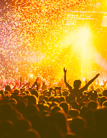 A crowded concert hall with scene stage orange and yellow lights, rock show performance, with people silhouette, colourful confetti explosion fired on dance floor air during concert festival