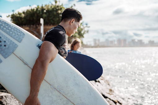 Profile view of a young and fit male surfer of Hawaiian and Japanese descent navigating a rocky shoreline while carrying his surfboard to the ocean with a view of Honolulu, Hawaii visible in the background.