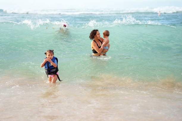 An Eurasian woman stands in the ocean near the shore and holds her one year old son as an ocean wave rushes near her and her three year old daughter who is wearing a lifejacket.