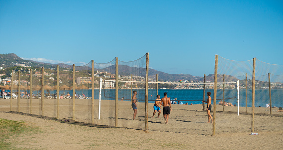 Young men playing Beach Volleyball on a netted-off court on Malagueta Beach, Málaga, Spain on a warm, sunny Saturday in late October. Other people are sunbathing on sunloungers further back in the photo.