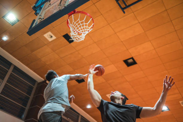 Low angle view on diverse basketball players jumping in the air to grab a rebound. Playing one on one basketball game in a sports hall.