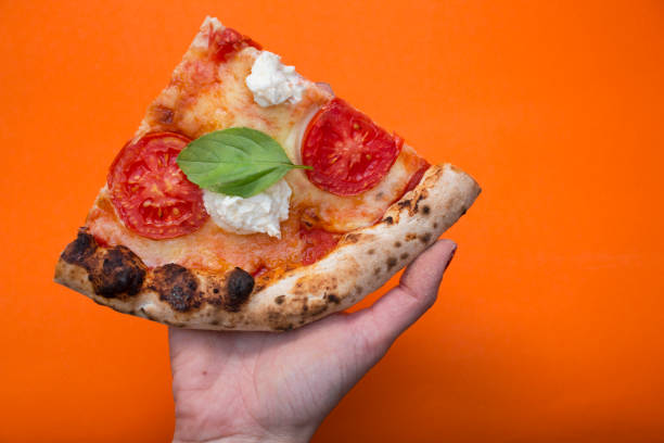 istock hand holding a slice of pizza with cheese, tomatoes and basil, orange background 1808652381