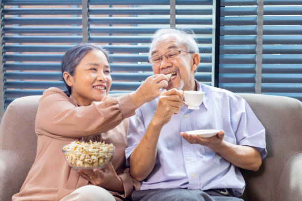 istock Senior asian couple is smiling while sitting on the sofa couch and having fun watching television with popcorn as snack for good mental health and longevity concept 1808805523