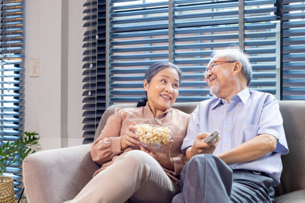 istock Senior asian couple is smiling while sitting on the sofa couch and having fun watching television with popcorn as snack for good mental health and longevity concept 1808805527