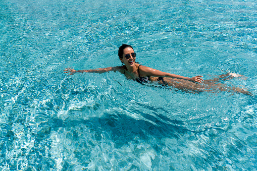 A female swimmer in a trendy bikini and sunglasses, swim in the crystal-clear water of a swimming pool on a sunny day.