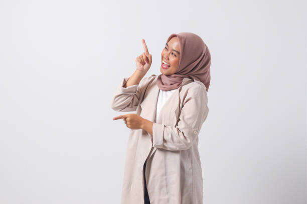Portrait of excited Asian hijab woman in casual suit pointing and showing product in her side with finger. Businesswoman concept. Isolated image on white background stock photo