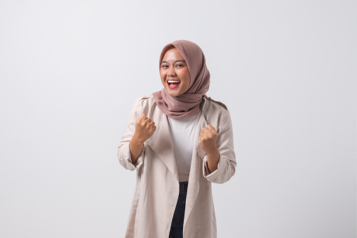 Portrait of excited Asian hijab woman in casual suit raising her fist, celebrating success. Businesswoman concept. Isolated image on white background