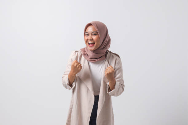 Portrait of excited Asian hijab woman in casual suit raising her fist, celebrating success. Businesswoman concept. Isolated image on white background stock photo