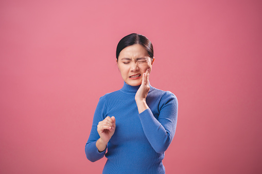 Asian woman was sick with toothache touching her cheek and standing isolated on pink background.