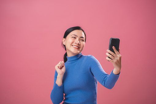 Asian woman happy enjoyed using smartphone for video call or take a photo standing isolated over pink background.