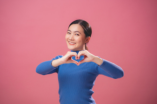 Asian woman falling in love happy smiling and making love symbol standing isolated on pink background.
