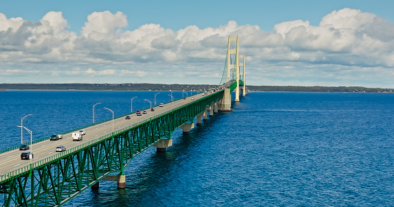 Aerial still image of Mackinac Bridge under a cloudy sky, with clear blue water underneath.