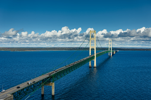 Aerial still image of Mackinac Bridge under a cloudy sky, with clear blue water underneath.