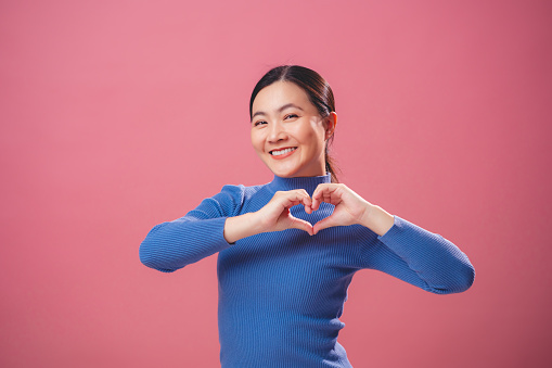 Asian woman falling in love happy smiling and making love symbol standing isolated on pink background.