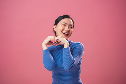 Asian woman falling in love happy smiling and making love symbol standing isolated on pink background.