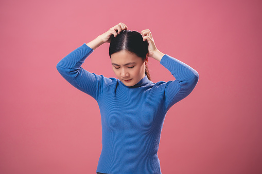 Asian woman bored and annoyed, scratching her head, standing isolated on pink background.