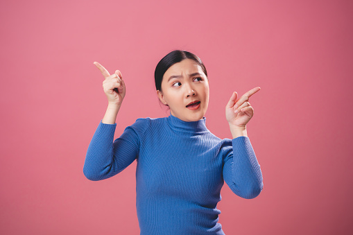 Asian woman thinking about problem, worried and confused pointing in different sides standing isolated on pink background.