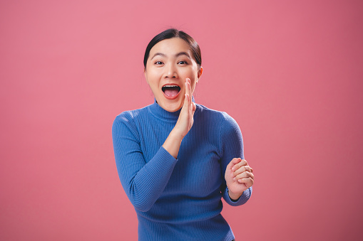Asian woman happy smiling making a whispering gesture telling secret or sharing news standing isolated on pink background.