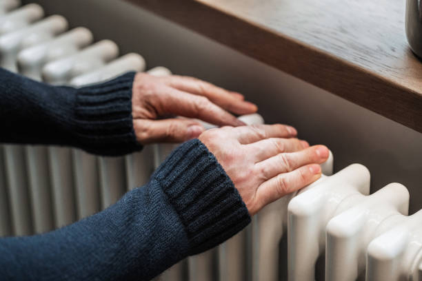 cold at home. a man tries to warm his hands on a radiator - huisverwarming fotos stockfoto's en -beelden