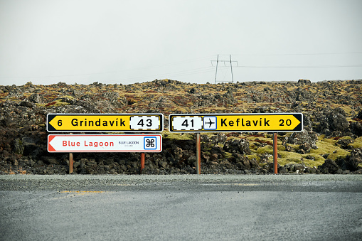 A road sign in Iceland guiding people towards Grindavik, Blue Lagoon, and Keflavik with lava, and moss in the background, in the middle of nature, and with an asphalt road in front of it.