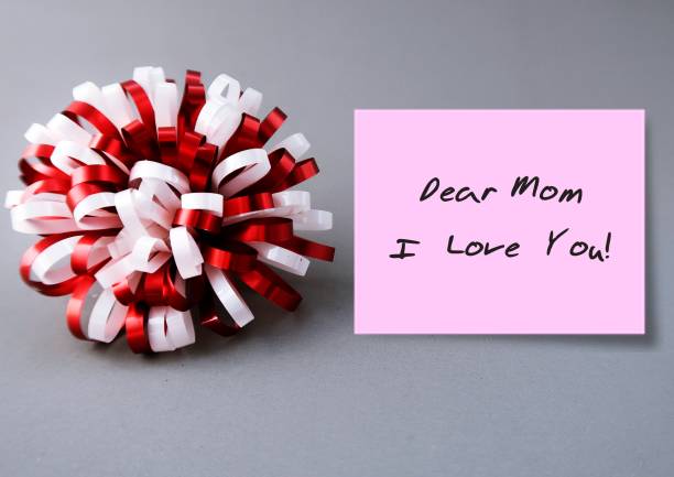 Gift bow and pink note on gray background with handwritten text DEAR MOM I LOVE YOU, concept of Happy Mother's Day card, honoring mother or to thank for all she's done and wish mom happiness