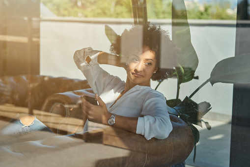 Through window of young ethnic female with Afro hairstyle relaxing in comfy armchair and looking away while browsing mobile phone in sunny day