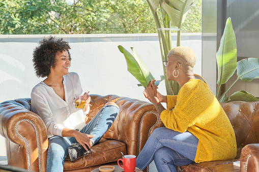 Young diverse female friends in casual clothes sitting on sofa and drinking beverages during conversation at home