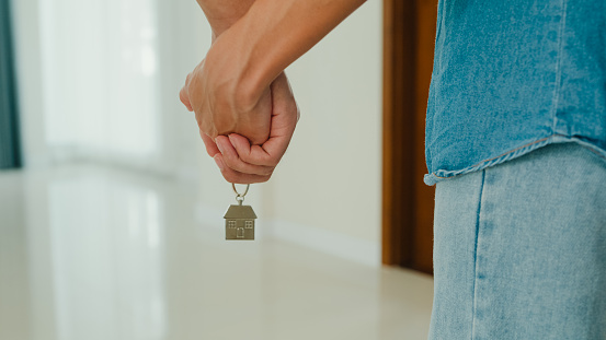 Young happy Asian couple hold hand and keychain inside new house in living room. Lifestyle spend time in house concept.