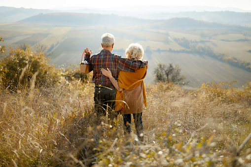 Back view of a senior couple standing embraced in tall grass on a hill and looking at view. Copy space.