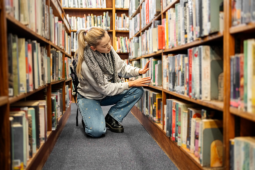 Young female student crouching by the lower shelves and looking for a book in university library