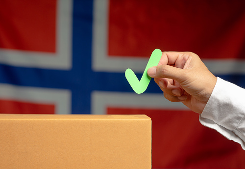Elections in Norway. Hand holding a green check mark symbol overhead the voting box at place election with the Norway flag background. Campaign to exercise the right to vote